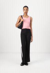 Débardeur rose, pantalon noir à jambes larges avec des rayures sur les côtés, sac à main noir et chaussures noires. Le modèle se tient devant un fond blanc uni.
