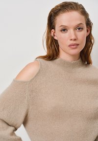 Beige gebreide trui met een hoge hals, uitsparingen bij de schouders en een zachte, fuzzy textuur. Eenvoudig, modern ontwerp met minimale details.