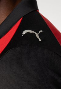 Ingrandimento di una maglia sportiva nera con un colletto rosso e un logo Puma argento sul lato sinistro in alto. Tessuto testurizzato visibile.