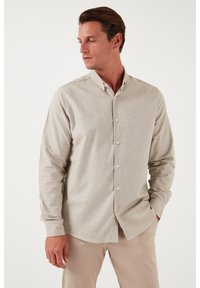 Camicia grigia chiara con maniche lunghe, colletto alzato, tessuto leggermente strutturato e chiusura frontale con bottoni. Il modello indossa pantaloni neutri.