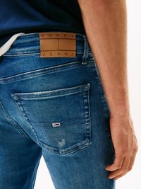 Plave traperice s srednjim pranjem, s smešnim kožnim oznakom "TOMMY JEANS" i malim crvenim, bijelim i plavim logotipom na stražnjem džepu.