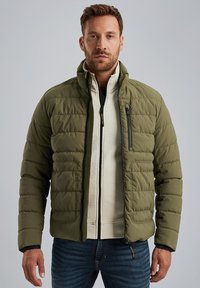 Olive green puffer jacket with a zippered chest pocket, quilted texture, and black accents, layered over a cream patterned sweater.