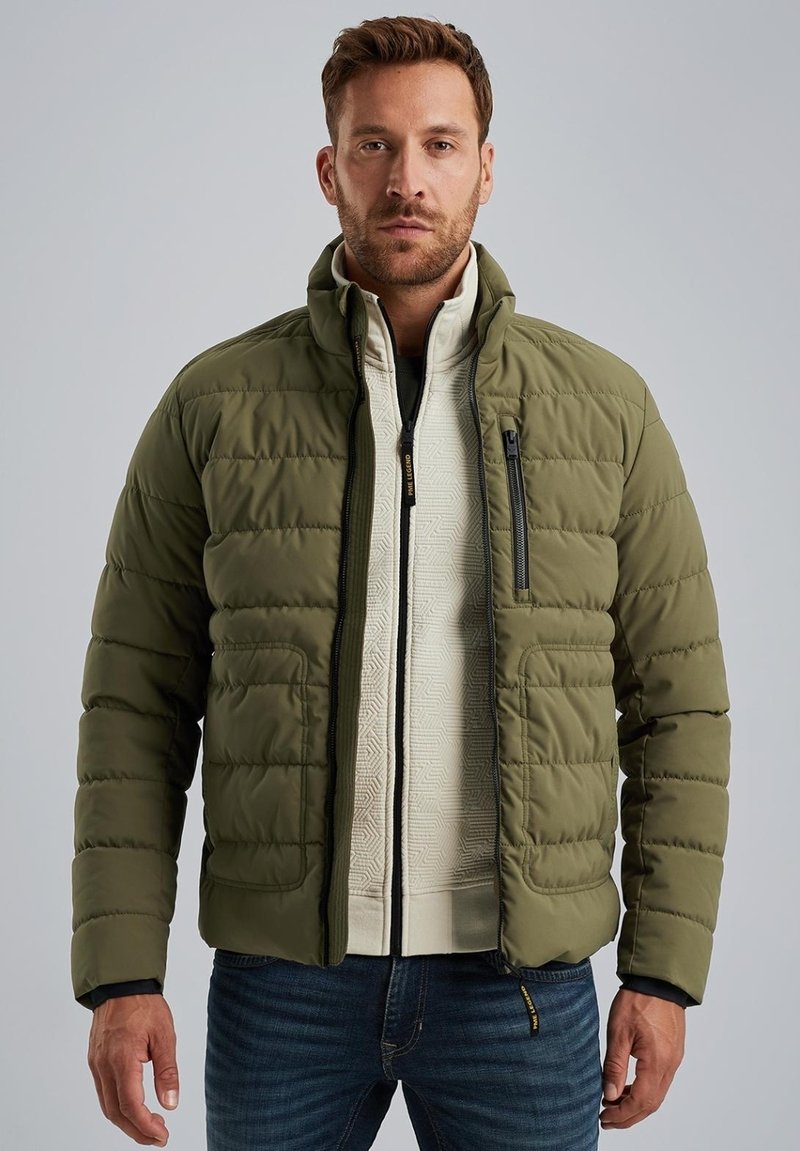 Olive green puffer jacket with a zippered chest pocket, quilted texture, and black accents, layered over a cream patterned sweater.