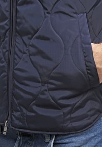 Chaqueta acolchada azul marino con una textura suave, patrón de costura curva, cierre de cremallera y detalle de bolsillo lateral.