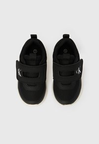 Ténis pretos para crianças com solas brancas, apresentando tecido de malha e uma tira de Velcro com o logótipo "cK" num fundo branco simples.