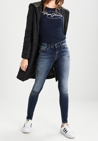 Zwarte gewatteerde jas, marineblauw shirt met wit logo, versleten blauwe skinny jeans en witte sneakers met zwarte strepen.