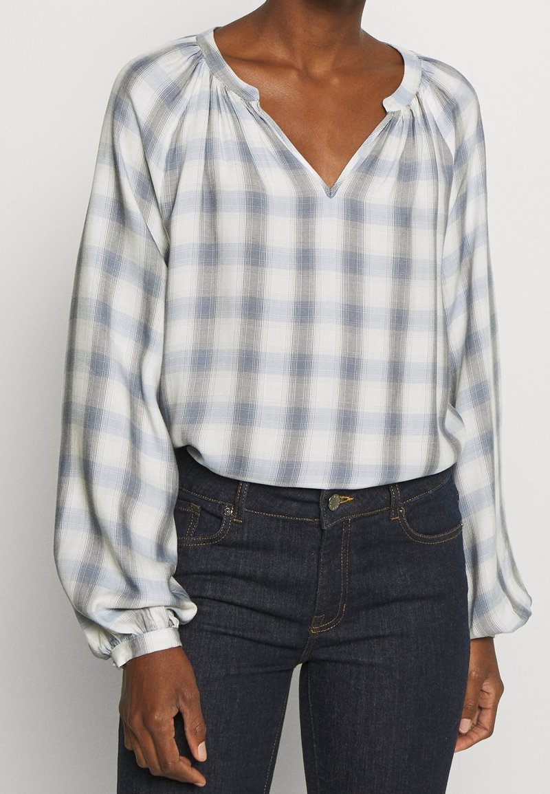 Blouse légère à manches longues, à carreaux bleu et blanc, avec un col en V et des coutures d'épaule froncées. Associée à un jean en denim foncé.