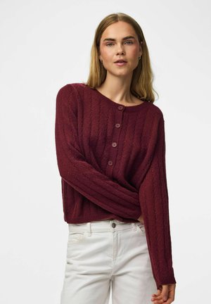 Femme portant un pull en maille bordeaux boutonné et un pantalon blanc, debout devant un fond clair uni.