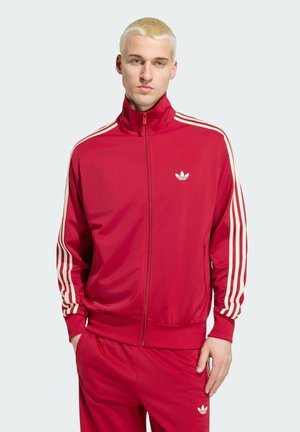 Jonge man met kort blond haar die een rode Adidas trainingspak met witte strepen en logo draagt, handen in de zakken, neutrale achtergrond.
