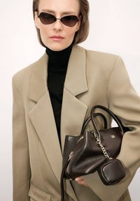 Ruskea nahkalaukku, jossa on teksturoitu pinta ja irrotettava pienempi pussukka, pidetään vaaleanbeigeä ylisuuri bleiseriä ja mustaa poolopaitaa vasten.