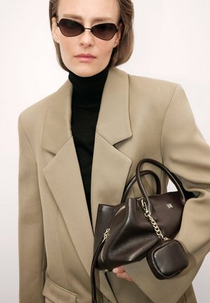 Brązowa skórzana torebka z fakturowaną powierzchnią i odczepianą mniejszą kieszonką, trzymana w zestawie z jasno beżową, oversize'ową marynarką i czarnym golfem.