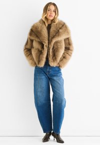 Brown faux fur jacket with a wide collar, worn over a brown top, paired with light blue high-waisted jeans and patterned heeled boots.