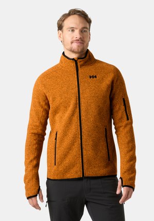 Man draagt een oranje fleecejack met ritssluiting en duimgaten, staand tegen een effen lichte achtergrond.