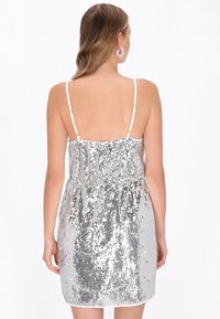 Srebrna mini sukienka z cekinami, z przezroczystym tyłem, regulowanymi cienkimi ramiączkami i elastycznym pasem. Teksturowana i refleksyjna powierzchnia.