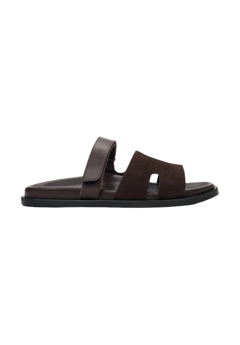 Brown suede slide sandal featuring a split design, adjustable strap, and a textured insole, complemented by a black rubber sole.