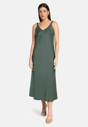 Frau steht und trägt ein ärmelloses, dunkelgrünes Midikleid mit dünnen Trägern sowie beige offene Zehenabsätze vor einem schlichten Hintergrund.