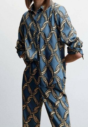 Žena v modré blůze a kalhotách s béžovým a černým propleteným řetězcem, rukávy vyhrnuté a ruce v kapsách.