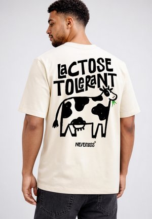 Man wearing cream t-shirt with black graphic cow and text "Lactose Tolerant" on the back, paired with black jeans.