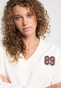 Valkoinen ylisuurikokoinen v-pääntiehuppari, jossa on teksturoitu kangas ja maroonivärinen brodeerattu "89"-patch vasemmassa rintapieluksessa.