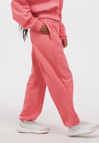 Mäkké ružové tepláky s elastickým pásom, bočnými vreckami a voľným strihom, skombinované s bielymi teniskami na jednoduchom pozadí.