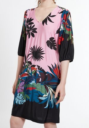 Femme portant une robe rose à décolleté en V avec des motifs de feuilles tropicales noires, bleues, rouges et vertes et des manches bouffantes noires trois-quarts.
