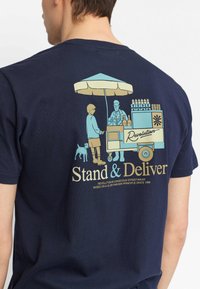 Marineblauw T-shirt met korte mouwen, met een kleurrafische afbeelding van een straatverkoper kar met een luifel, mensen en een hond. De tekst luidt "Stand & Deliver."