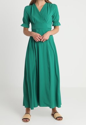Robe portefeuille verte avec des manches bouffantes, fabriquée dans un tissu lisse. Design long jusqu'au sol, avec une taille cintrée et une jupe fluide. Portée avec des sandales plates.
