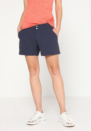 Woman wearing navy blue shorts, coral sleeveless top, and white woven sneakers, standing with hands in pockets against a plain background.