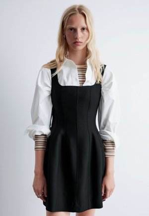 Schwarzes, tailliertes Kleid mit strukturierten Paneelen, kombiniert mit einem weißen Hemd mit gestreiften Ärmeln. Glatte Stofftextur, minimalistisches Design.