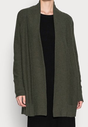 Persona che indossa un cardigan a coste verde scuro aperto sopra un vestito a coste nero, in piedi davanti a uno sfondo chiaro e uniforme.