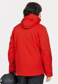 Rote wasserdichte Jacke mit Kapuze, die eine glatte Textur und ein schlichtes Design aufweist. Die Person hält einen schwarzen Helm in der Hand.