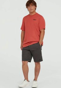 Young man wearing a red O'Neill t-shirt, dark gray shorts, and white sneakers, standing with one hand in pocket.