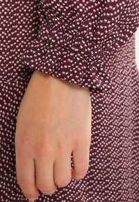 Bordeauxklänning med ett texturerat mönster av små hjärtan och en volangdetalj på lång ärm, som visar en hand i vila.