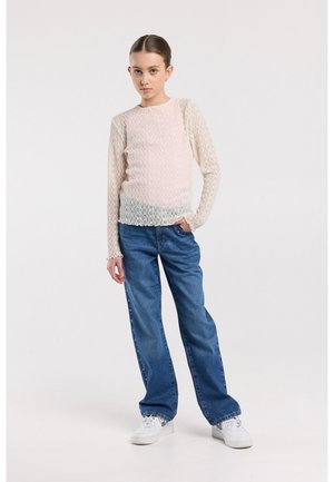 Jonge persoon die staat met de hand in de zak, gekleed in een doorschijnend shirt met lange mouwen, blauwe spijkerbroek met zijdetails en witte sportschoenen, tegen een effen achtergrond.