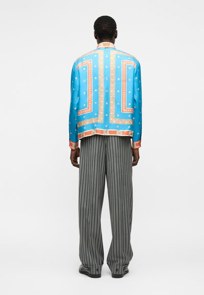 Muž oblečený v jasne modrej vzorovanej dlhorukávovej košeli a čierno-bielych širokých nohaviciach s vertikálnymi pruhmi, stojaci čelom odvrátený.