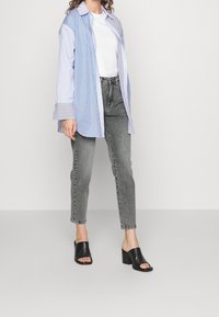 Kvinna iförd en lös blårandig skjorta över en vit t-shirt, grå jeans med hög midja och svarta klackskor med öppen tå, stående på grått golv.