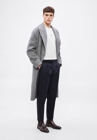 Γκρι παλτό από μάλλινο ύφασμα ψαροκόκαλο, λευκό μπλουζάκι, μπλε ναυτικό παντελόνι με πιέτες και καφέ δερμάτινα μποτάκια μέχρι τον αστράγαλο. Το μοντέλο στέκεται με τα χέρια στις τσέπες.