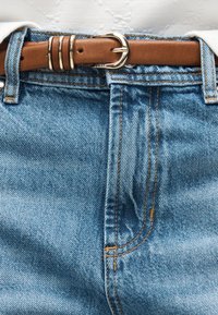 Καφέ δερμάτινη ζώνη με ασημένια πόρπη στερεωμένη μέσα από τις θηλιές της ζώνης σε μπλε τζιν τζιν με ορατά ραφές.