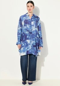 Abito camicia blu tie-dye con abbottonatura frontale, maniche lunghe e orlo curvo, abbinato a pantaloni larghi in denim scuro.