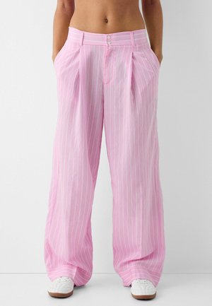 Frau trägt hochgeschnittene, weit geschnittene rosa Hosen mit weißen vertikalen Streifen und weißen Sneakers, steht mit den Händen in den Taschen.