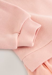 Világos rózsaszín pulóver anyag lágy tapintással; bordázott mandzsettákkal és szegéllyel, rétegezett kialakítással, sima és pliszírozott felületekkel.