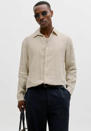 Mann iført beige knappet skjorte, mørke bukser, solbriller, holder en svart veske med den ene hånden i lommen mot en ensfarget lys bakgrunn.