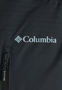 Tessuto nero con una trama a griglia che presenta un logo Columbia in azzurro chiaro e una cerniera nera chiusa sul lato sinistro.