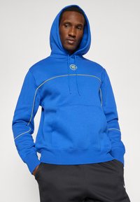 Sudadera con capucha azul hecha de una mezcla de algodón. Presenta acentos de costura amarilla y un bolsillo tipo canguro. Modelo con pantalones negros.