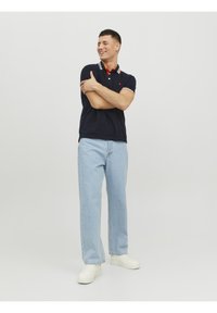 Marineblå polo skjorte med kontrasterende rød og hvid krave, parret med lyseblå lige ben jeans og hvide sneakers. Enkel baggrund.