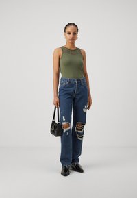 Olivengrønn tanktop med blonde detaljer ved halsen, sammen med slitte blå denimjeans med store hull, og svarte sko.