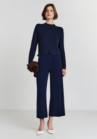 Navy gestrickter Kabelpullover, cropped Design, kombiniert mit weit geschnittenen navyfarbigen Hosen. Das Model hält ein braunes Stoffaccessoire und trägt weiße High Heels.