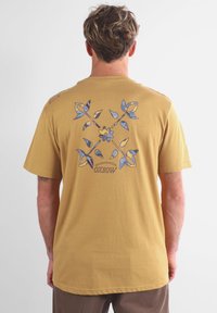 T-shirt en coton jaune avec un graphique floral bleu au dos, manches courtes et ourlet arrondi. Marque Oxbow sur l'épaule.