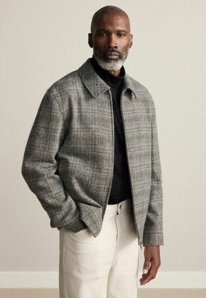 Graue Karomuster-Zip-Jacke mit einem kragenförmigen Halsauschnitt, strukturiertem Stoff und Fronttaschen, kombiniert mit einem schwarzen Shirt und cremefarbigen Hosen.