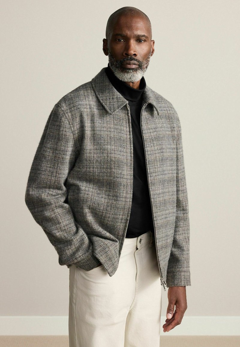 Gray plaid zip-up jacket with a collared neckline, textured fabric, and front pockets, paired with a black shirt and cream-colored trousers.
